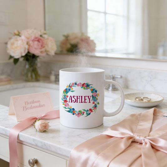 a white mug with a floral design, a pink robe, a card with the name 'Ashley', and a vase of pink flowers on a marble countertop.
