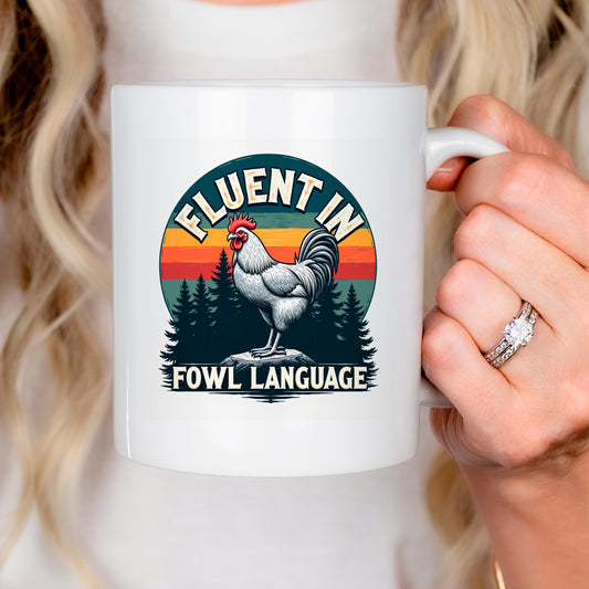 A person is holding a white mug with a graphic design featuring a rooster and the text "FLUENT IN FOWL LANGUAGE".