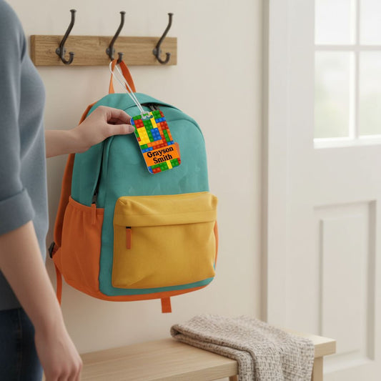A person is holding a colorful backpack with a name tag attached to it, standing in front of a wooden coat rack.