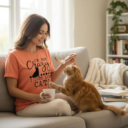 A woman in an orange t-shirt is sitting on a couch, feeding a cat while holding a cup of coffee.