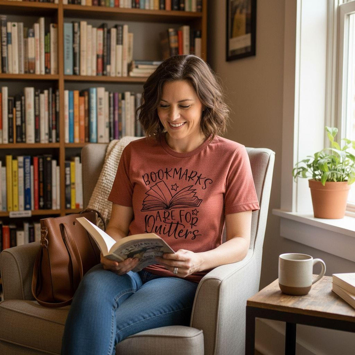 A woman is sitting in a chair, reading a book, and smiling. She is wearing a red t-shirt with the text "Bookmarks are for quilters" printed on it. The room has a bookshelf filled with books, and there is a potted plant and a coffee cup on a table nearby.