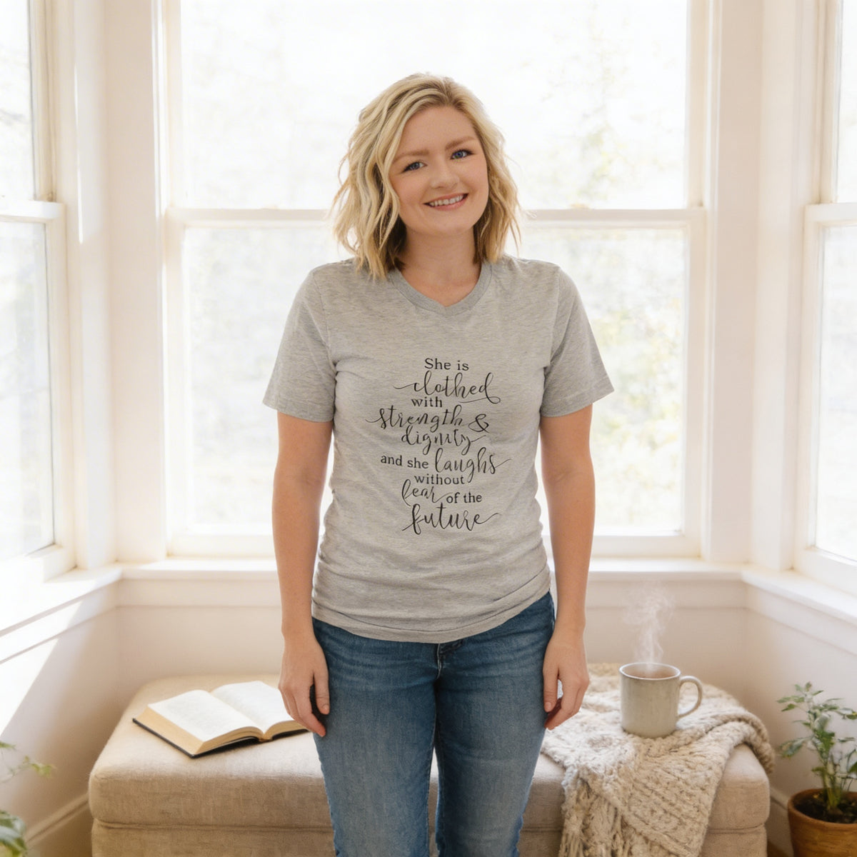 A woman wearing a gray t-shirt with text stands in front of a window, smiling and holding a book.
