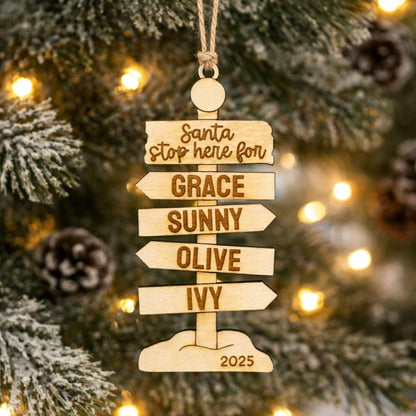 A wooden ornament hanging on a Christmas tree, featuring a directional sign with the names of the five daughters of Santa Claus.