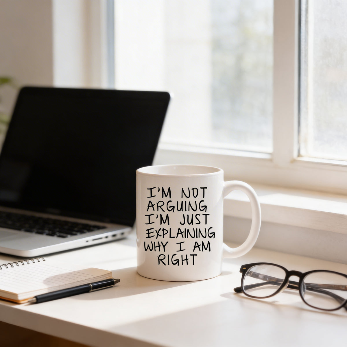 A white mug with black text sits on a desk next to a laptop, with a pair of glasses and a notebook nearby.