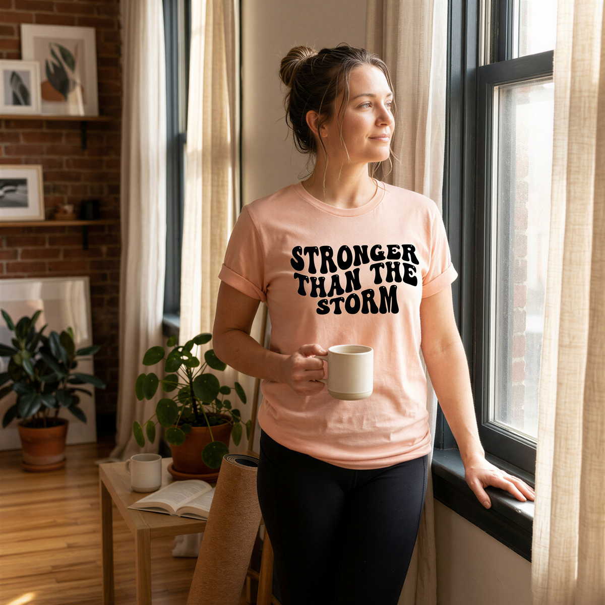A woman wearing a pink t-shirt with the text "STRONGER THAN THE STORM" is standing near a window, holding a coffee mug and looking out.