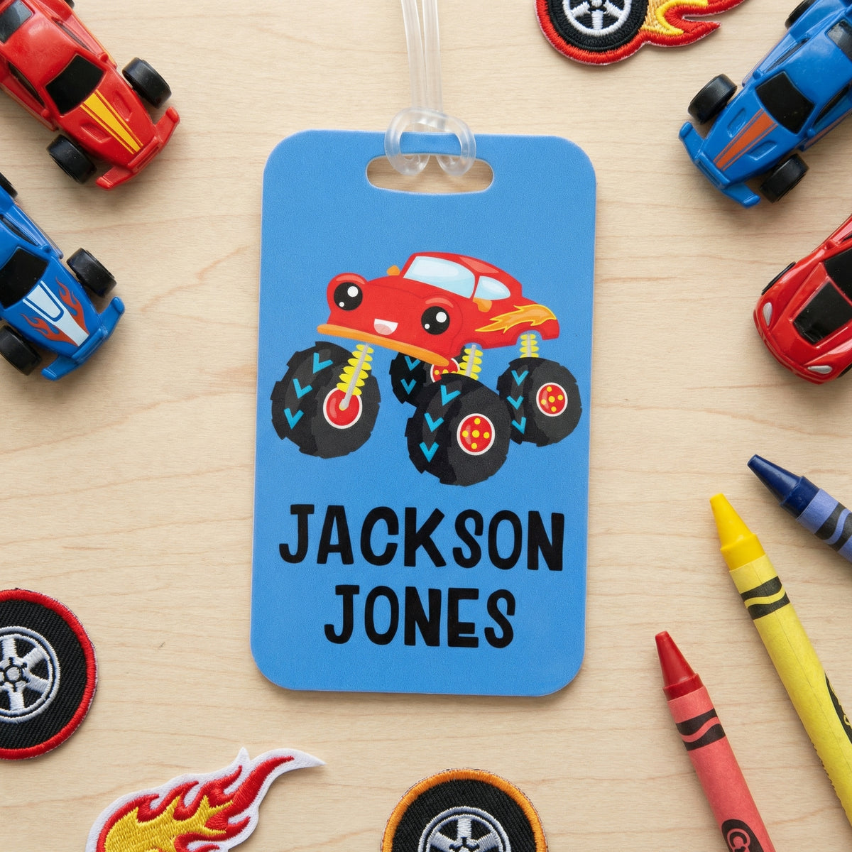 A blue luggage tag with a cartoon monster car design, surrounded by various children's toys and personal items on a wooden surface.