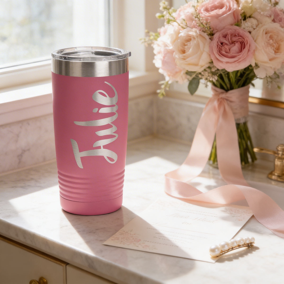 A pink tumbler with the name "Julie" printed on it, placed on a marble countertop next to a bouquet of pink and white flowers and a pearl hair clip.