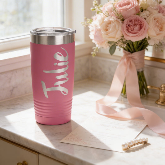 A pink tumbler with the name "Julie" printed on it, placed on a marble countertop next to a bouquet of pink and white flowers and a pearl hair clip.