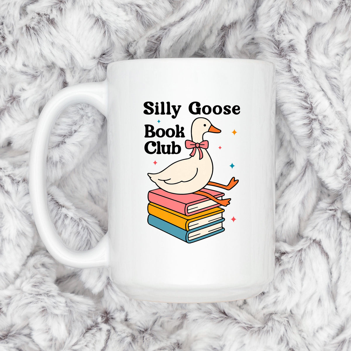 A white mug with a graphic design featuring a cartoon goose sitting on a stack of colorful books, with the text "Silly Goose Book Club" displayed above the goose.
