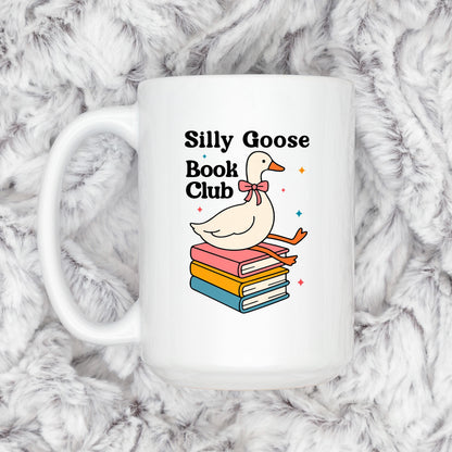 A white mug with a graphic design featuring a cartoon goose sitting on a stack of colorful books, with the text "Silly Goose Book Club" displayed above the goose.