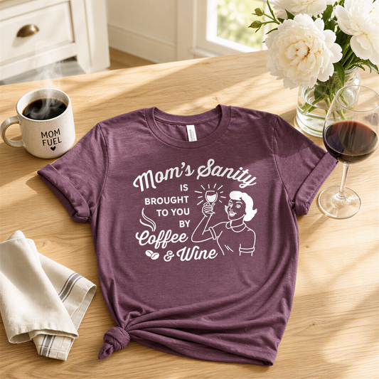 A purple t-shirt with a humorous text printed on it, along with a cup of coffee, a glass of red wine, and a vase of white flowers on a wooden table.