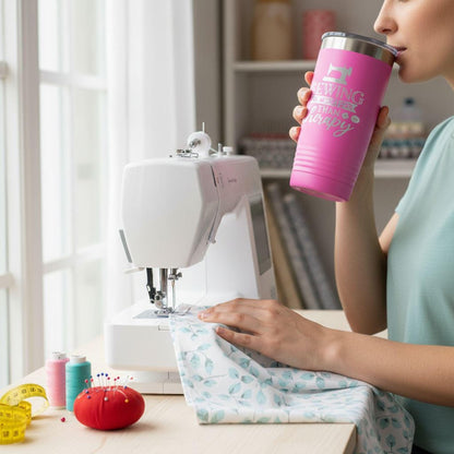 A person is working on a sewing machine, holding a pink tumbler with the text "Sewing is better than therapy" printed on it.