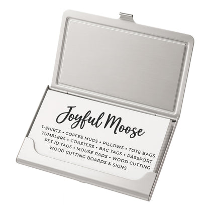 a business card holder with a business card inside. The card holder has a silver color and is open, revealing the card inside. The card contains text that provides information about the business, including the name 'Joyful Moose' and a list of items they offer, such as T-shirts, coffee mugs, pillows, tote bags, pet tags, coasters, and cutting boards.