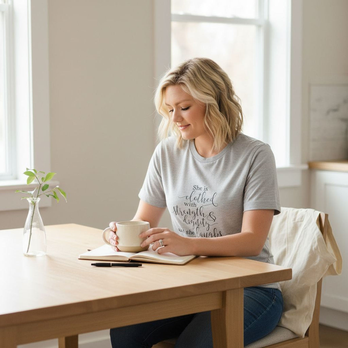 A woman sits at a wooden table, holding a mug and looking down at a notebook, with a vase of flowers and a plant nearby.