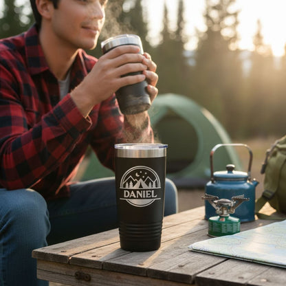 A man in a red and black plaid shirt is sitting at a wooden table, holding a black thermos with a logo on it, while a blue camping stove and a green backpack are also visible on the table.
