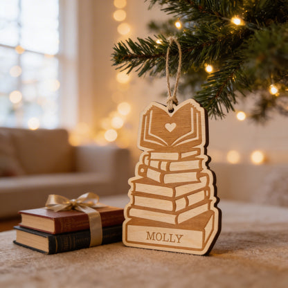 A wooden ornament shaped like a stack of books is hanging from a Christmas tree, with a gift box and a cozy setting visible in the background.