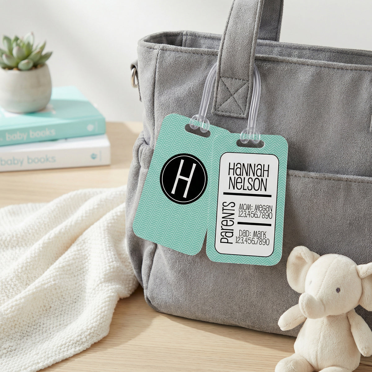A gray tote bag with a teal patterned tag attached, featuring a name tag for a patient named Hannah Nelson. The bag is placed on a wooden surface, with a white blanket and a stuffed elephant toy nearby.