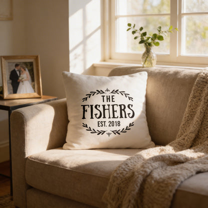 A white pillow with the text "THE FISHER'S EST. 2018" printed on it, sitting on a beige couch in a cozy living room. There is a framed photo of a couple on a nearby table, and a vase of greenery on the windowsill.