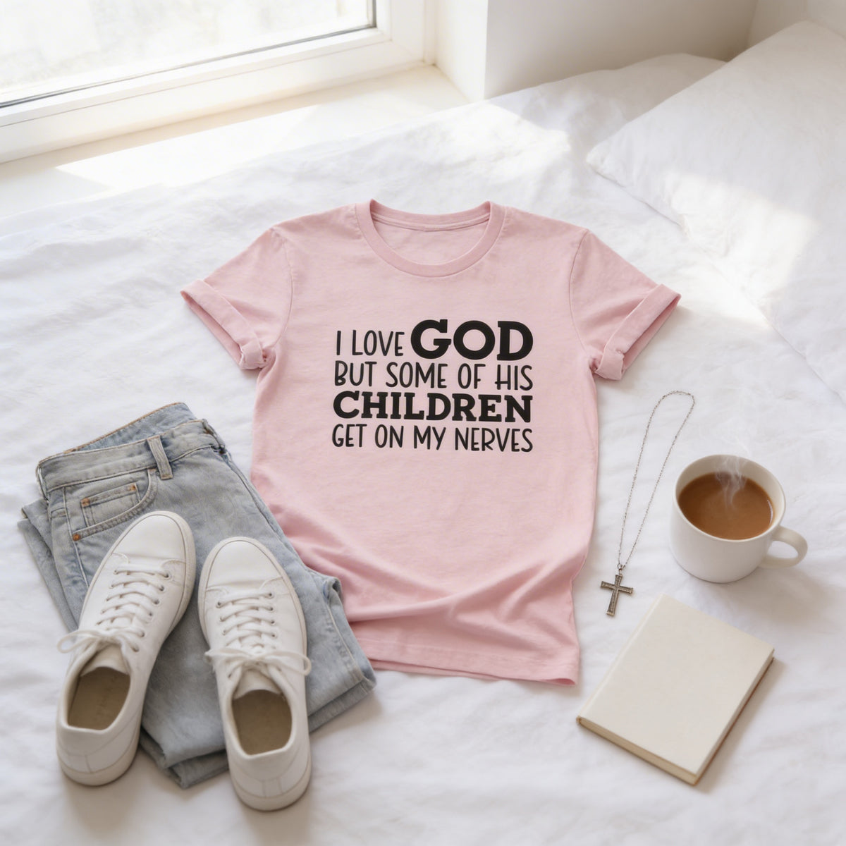 A pink t-shirt with text, a pair of jeans, white sneakers, a cross necklace, a mug of coffee, and a notebook are arranged on a bed.