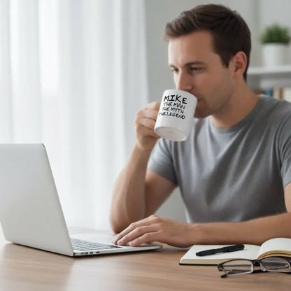 A man is sitting at a desk, working on a laptop while drinking from a mug that says "Mike, the man who will be the legend".