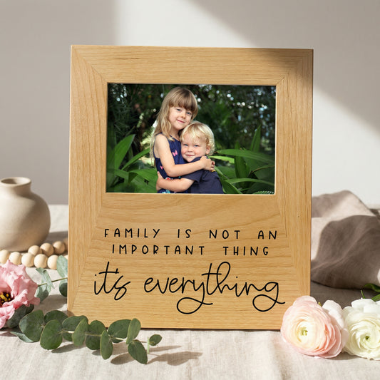 A wooden picture frame with a photo of two children inside, surrounded by flowers and greenery. The frame has a message that says "Family is not an important thing, it's everything".