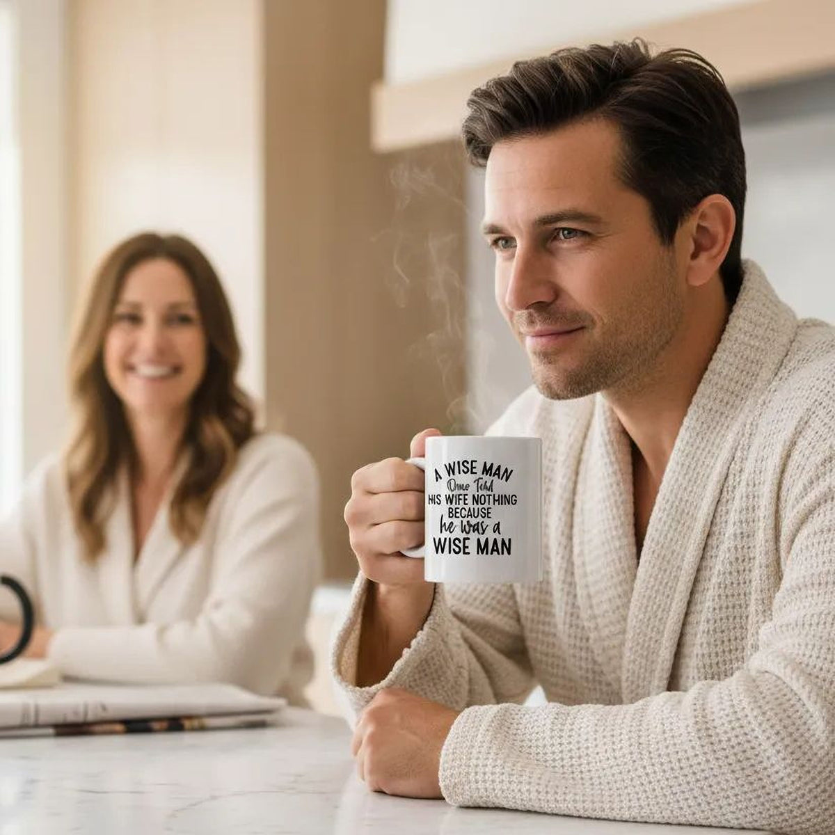 A man in a white robe is sitting at a table, holding a mug with text on it, while a woman is sitting across from him.