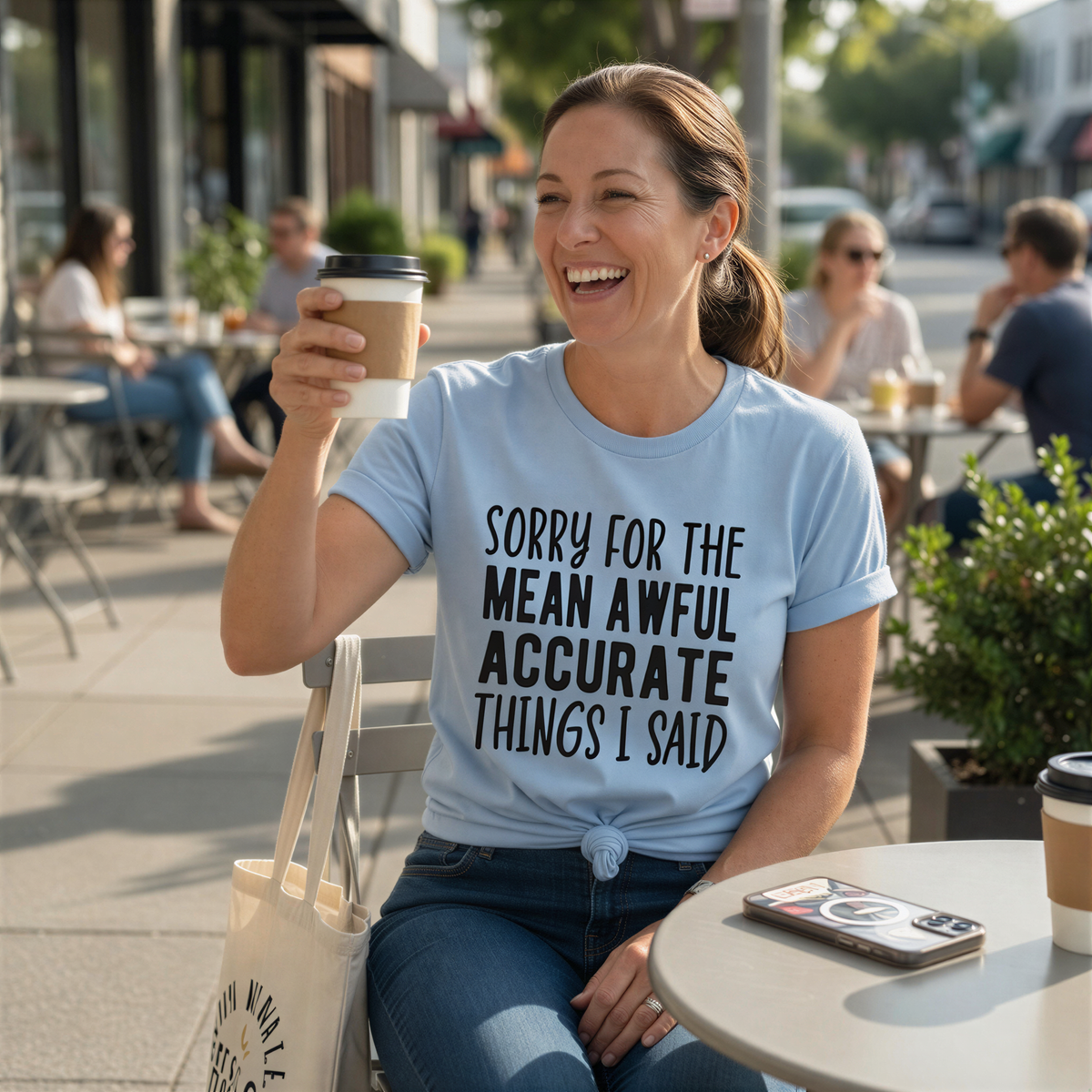 A woman wearing a light blue t-shirt with a humorous text is sitting at an outdoor table, smiling and holding a coffee cup.