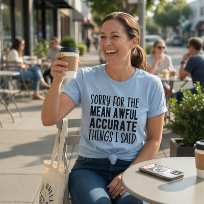 A woman wearing a light blue t-shirt with a humorous text is sitting at an outdoor table, smiling and holding a coffee cup.