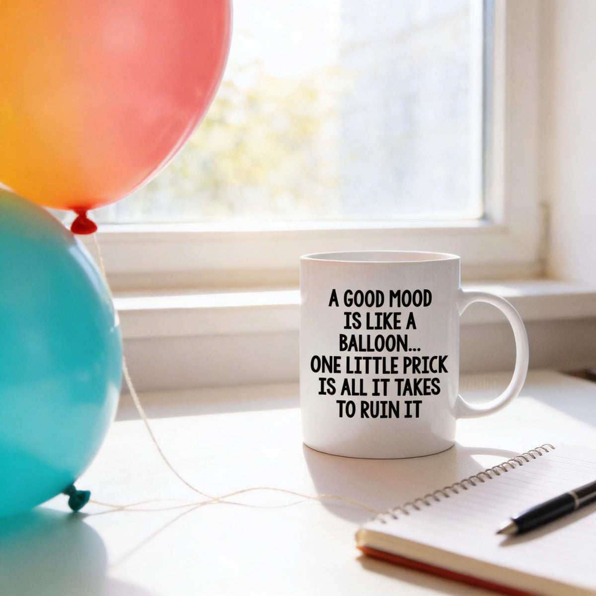 A white mug with a humorous text printed on it sits on a desk next to a notebook and a pen, with colorful balloons floating in the background.