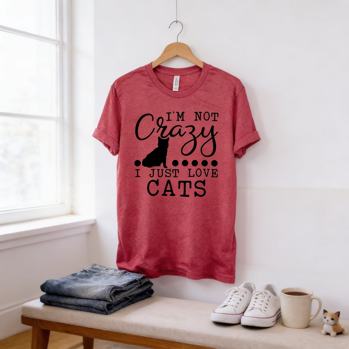 A red t-shirt with the text "I'M NOT CRAZY I JUST LOVE CATS" printed on it, along with a silhouette of a cat. The t-shirt is hanging on a wooden hanger, and there are a pair of jeans and a pair of white sneakers placed on a bench in the foreground.