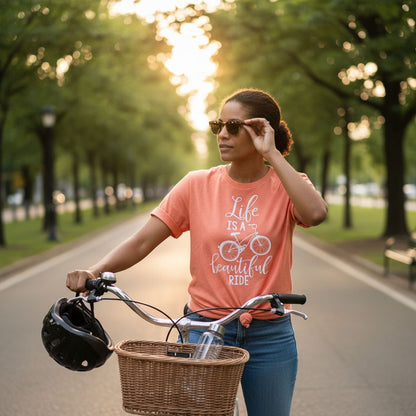 A woman wearing a pink t-shirt with a bicycle graphic and the text "Life is a beautiful ride" is standing next to her bicycle with a basket, wearing sunglasses and holding a helmet.