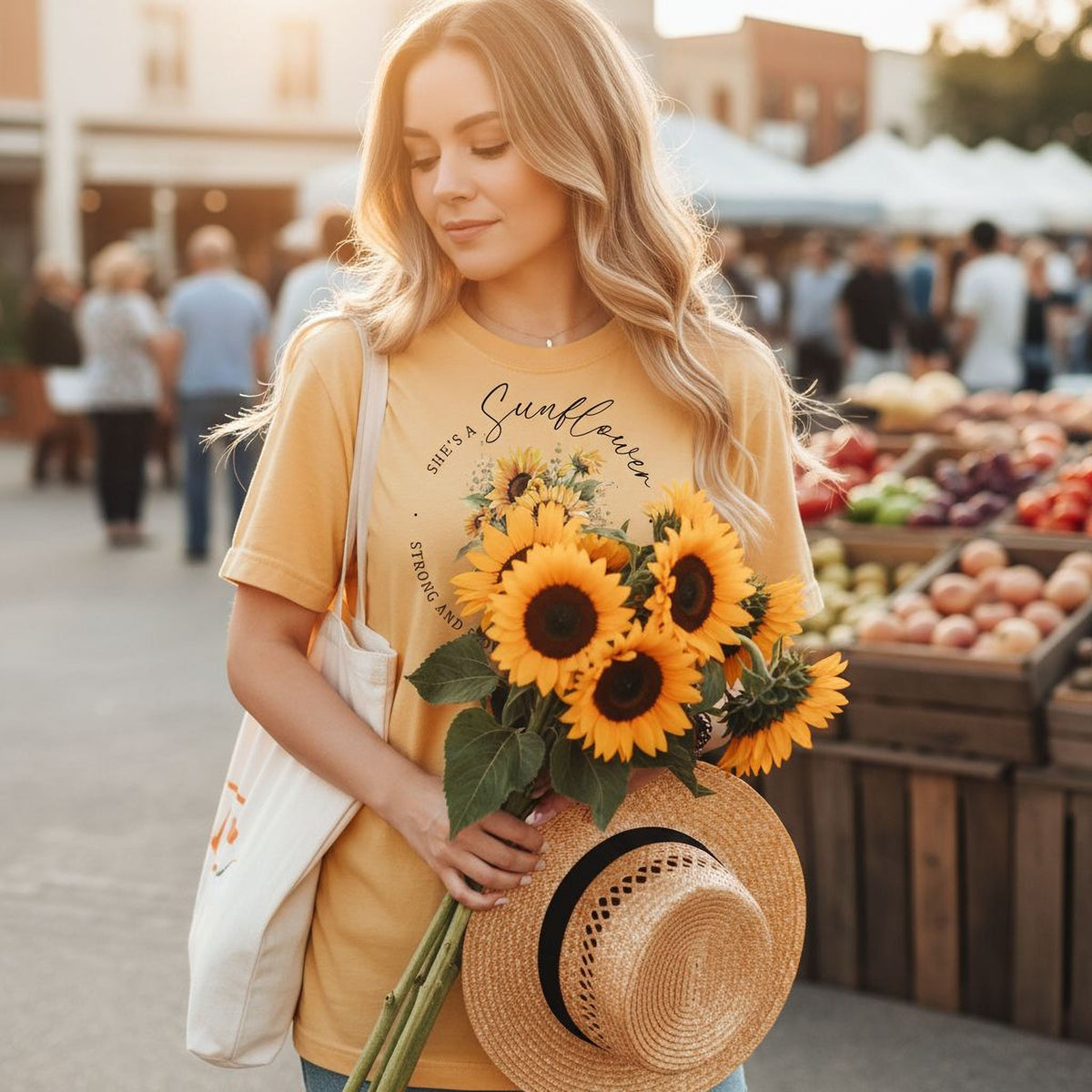 A woman in a yellow t-shirt holding a bouquet of sunflowers stands in front of a fruit stand at an outdoor market.
