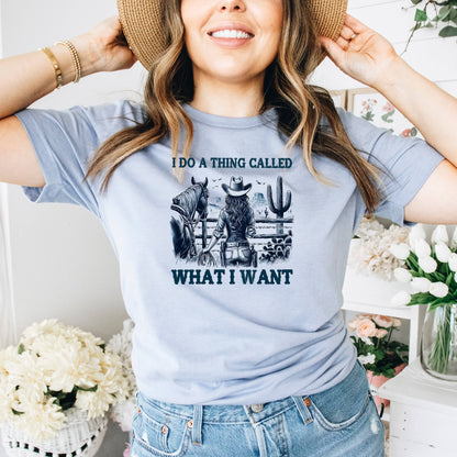 A woman wearing a blue t-shirt with a graphic design of a cowboy and a cactus, smiling and holding her hat up to her head.