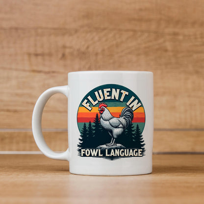 A white ceramic mug with a graphic design featuring a rooster and the text "FLUENT IN FOWL LANGUAGE" against a sunset background.