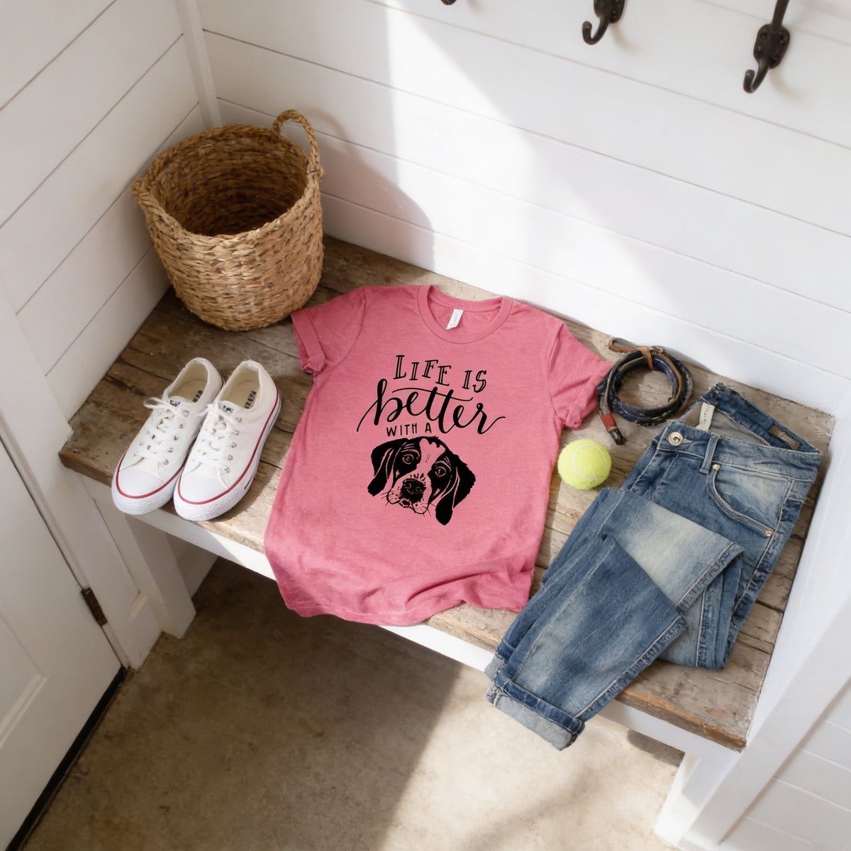 a pink t-shirt with a dog graphic, a pair of jeans, a pair of white sneakers, a woven basket, and a tennis ball on a wooden bench.