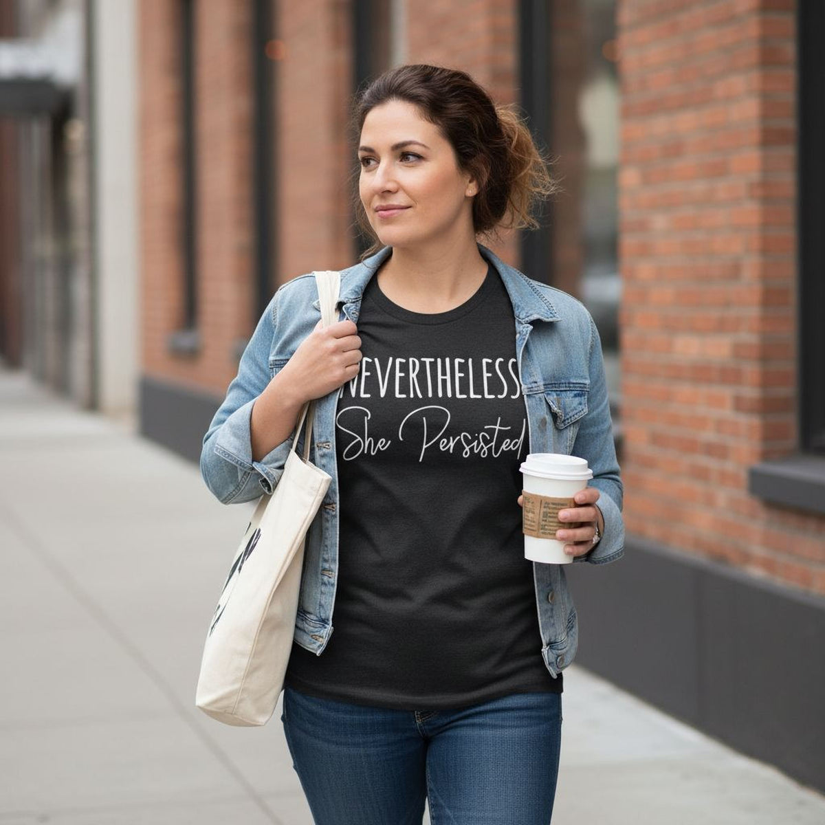 A woman wearing a denim jacket, carrying a white tote bag, and holding a coffee cup is walking on a sidewalk.