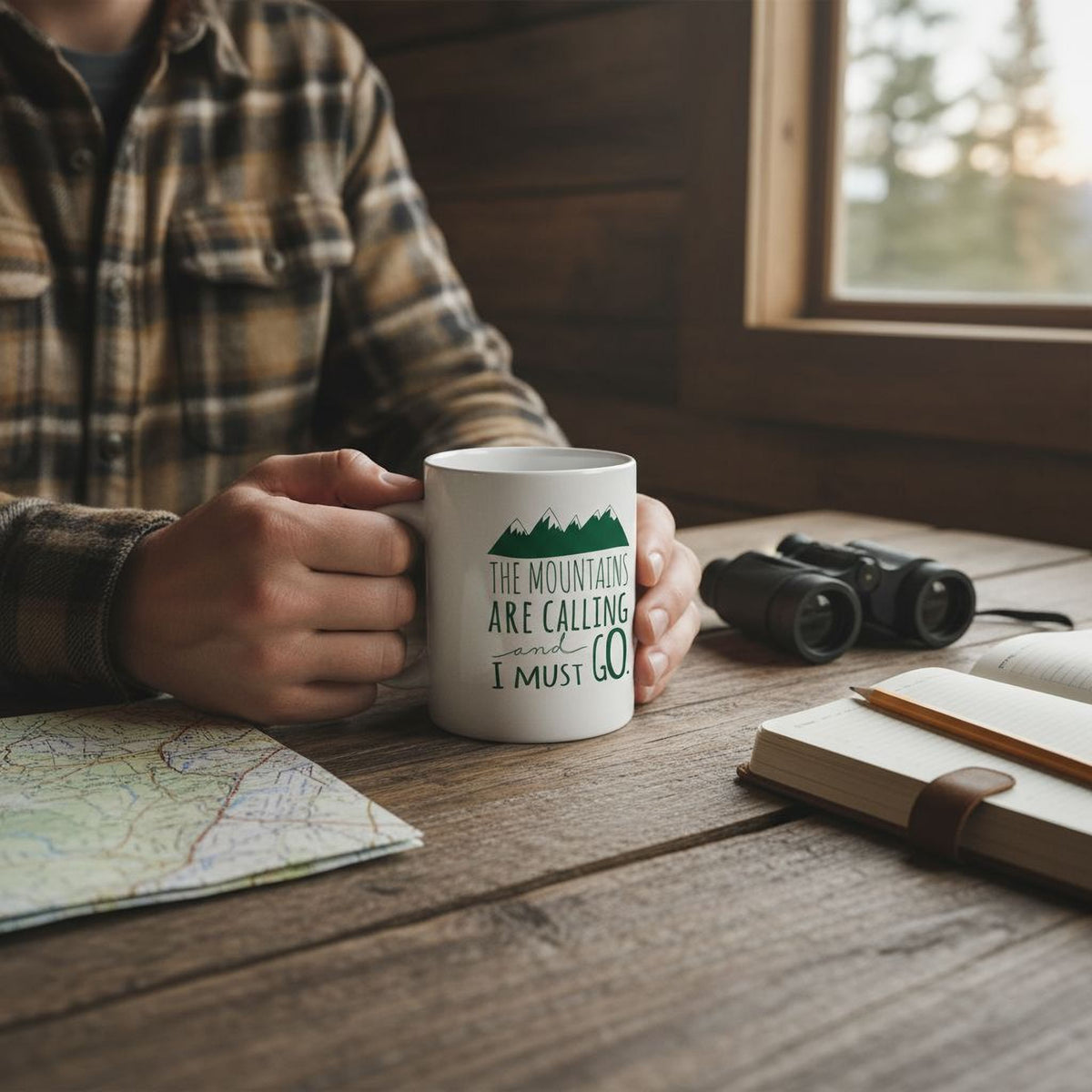 A person is sitting at a wooden table, holding a white mug with the text "The mountains are calling I must go" printed on it. The table also has a map, a notebook, and a pair of binoculars.