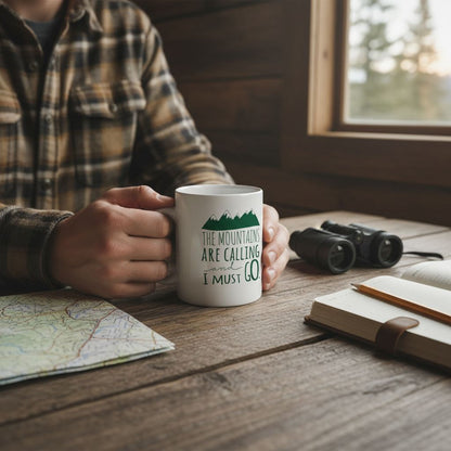 A person is sitting at a wooden table, holding a white mug with the text "The mountains are calling I must go" printed on it. The table also has a map, a notebook, and a pair of binoculars.