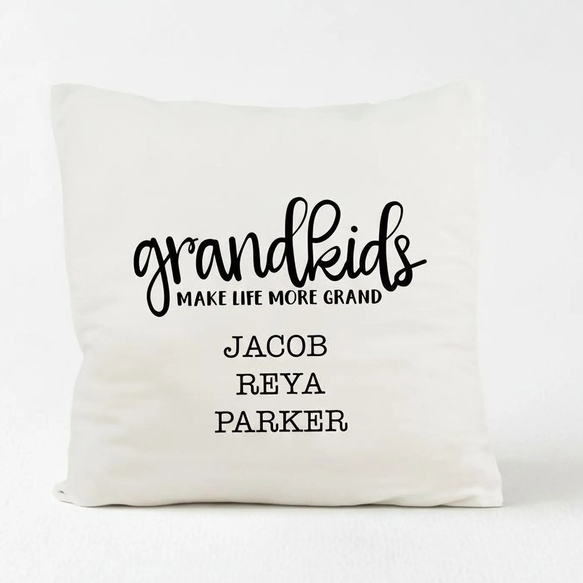 A white pillow with black text that says "grandkids make life more grand" and names "Jacob, Reya, Parker".