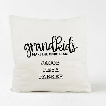 A white pillow with black text that says "grandkids make life more grand" and names "Jacob, Reya, Parker".