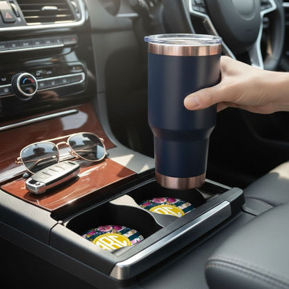 A person's hand is holding a blue thermos cup in the car's cup holder, with sunglasses and a key visible on the car's console.