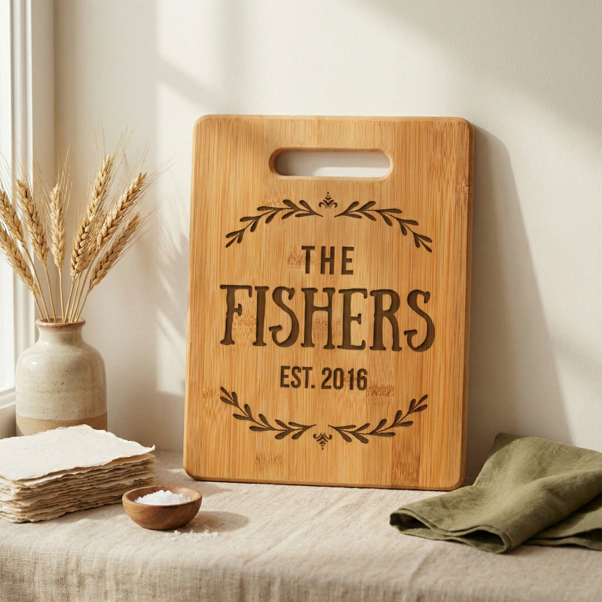 a wooden cutting board with the text "The Fishers EST. 2016" engraved on it, placed on a table next to a vase with wheat stalks and a folded green napkin.