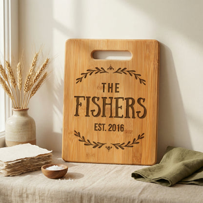 a wooden cutting board with the text "The Fishers EST. 2016" engraved on it, placed on a table next to a vase with wheat stalks and a folded green napkin.