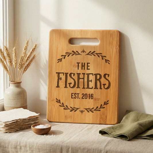 a wooden cutting board with the text "The Fishers EST. 2016" engraved on it, placed on a table next to a vase with wheat stalks and a folded green napkin.