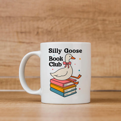 A white mug with a graphic design featuring a cartoon goose sitting on a stack of colorful books, with the text "Silly Goose Book Club" printed on it.