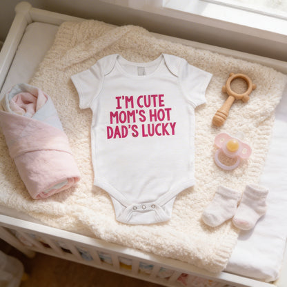 A baby's outfit, including a white onesie with the text "I'm CUTE MOM'S HOT DAD'S LUCKY" printed on it, is laid out on a white blanket. The outfit is accompanied by a pacifier, a stuffed animal, and a pair of socks.