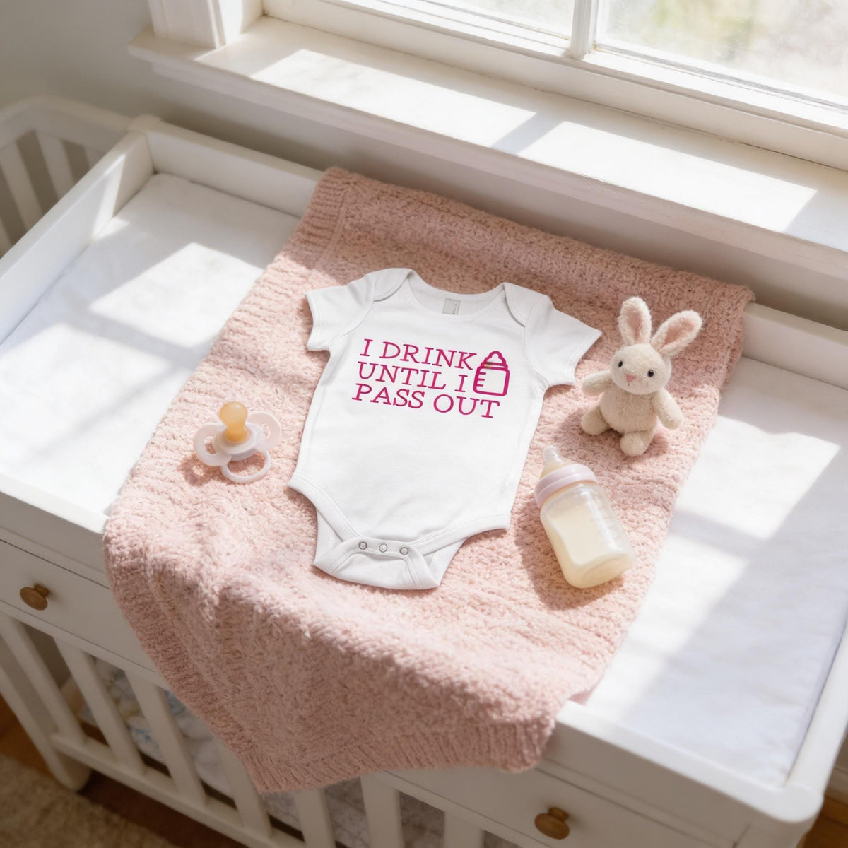 A baby's onesie with the text "I DRINK UNTIL I PASS OUT" is placed on a pink blanket next to a pacifier and a stuffed bunny.