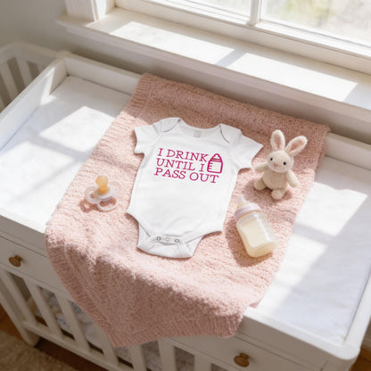 A baby's onesie with the text "I DRINK UNTIL I PASS OUT" is placed on a pink blanket next to a pacifier and a stuffed bunny.