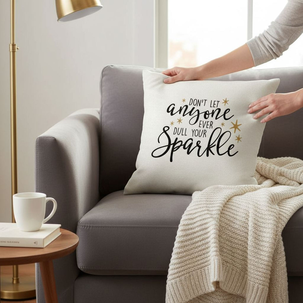 A person is holding a white pillow with black text that says "Don't let anyone dull your sparkle" in a cozy living room setting with a gray couch, a wooden side table, and a white mug.