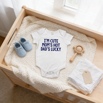 A baby's onesie with the text "I'M CUTE MOM'S HOT DAD'S LUCKY" is displayed on a wooden dresser, accompanied by a pair of blue baby shoes, a wooden rattle, and a white towel.